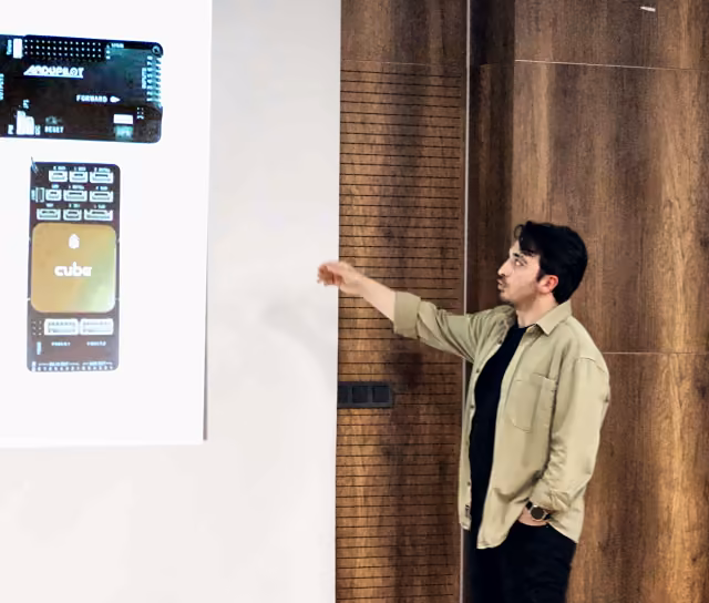 Furkan IŞIKAY Next'26 sahnesinde, perdedeki ArduPilot ve Cube uçuş kartlarını gösteriyor