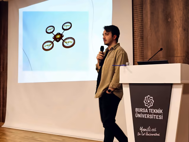 Furkan IŞIKAY Bursa Teknik Üniversitesi sahnesinde mikrofonla konuşuyor, arkasındaki perdede bir drone çizimi